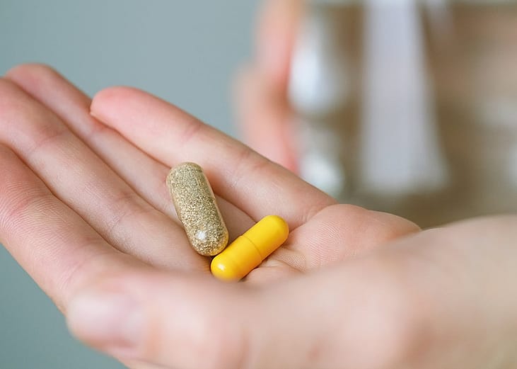 A hand with two capsules and a glass of water in the background.