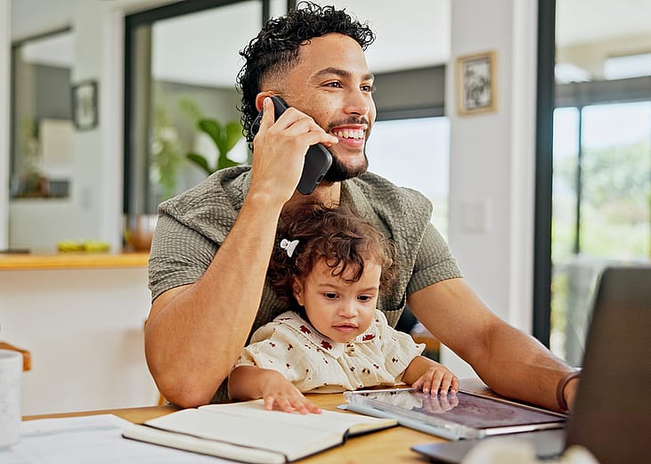 A happy young dad at home juggling calls, working, and looking after his young daughter on his lap.