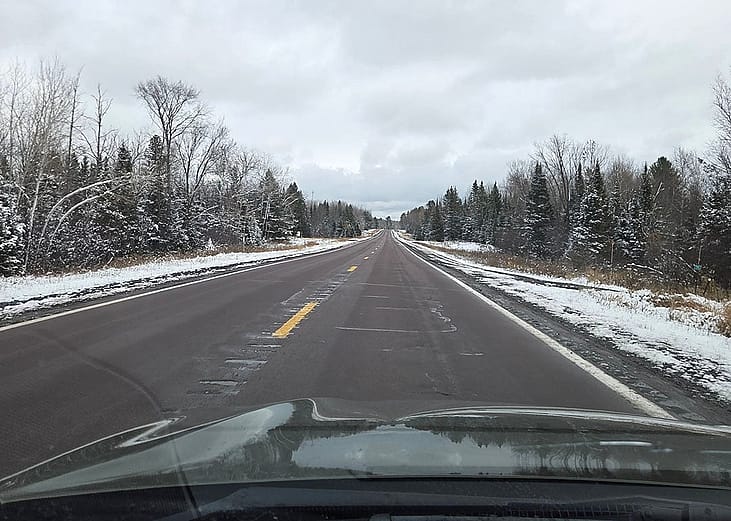 View of a highway surrounded by evergreen and bare trees with patches of snow.