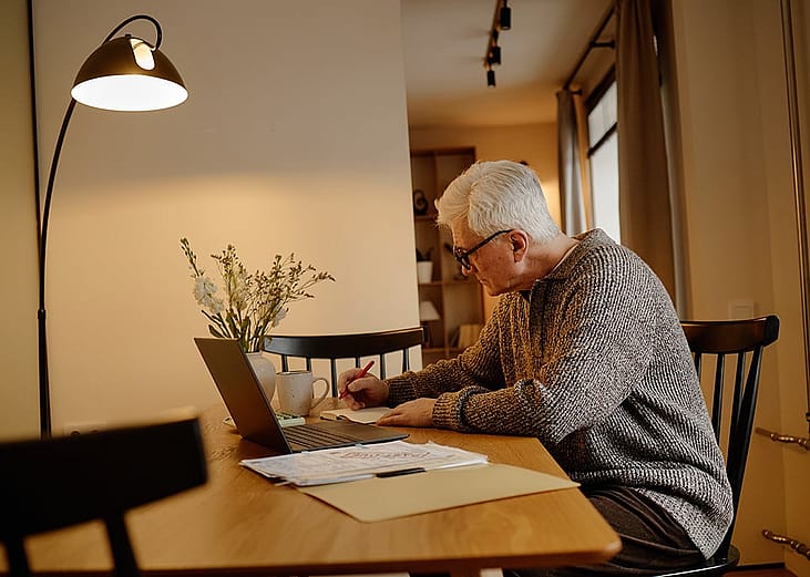 An elderly person writing notes, calculating bills, and using a laptop at home.