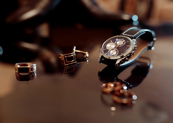 A luxury watch alongside a few rings on top of a glass table.