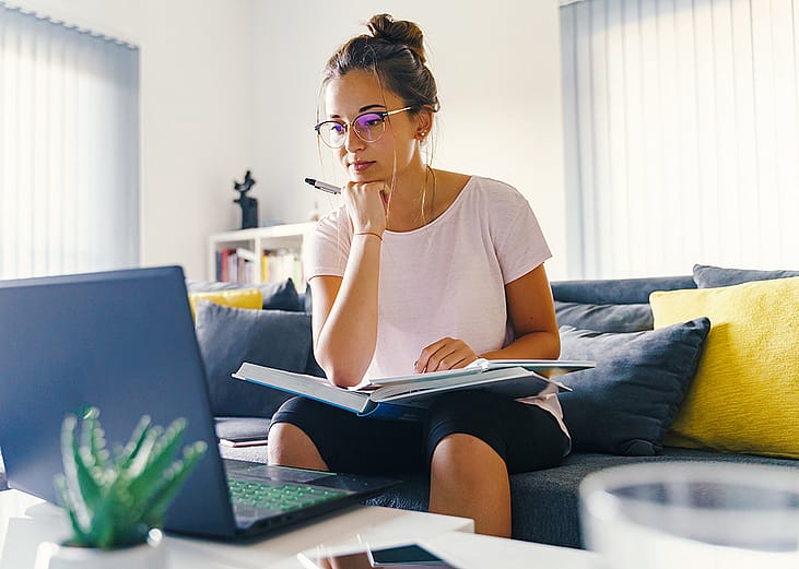A young woman at home reviewing information online and using a pen and notebook.
