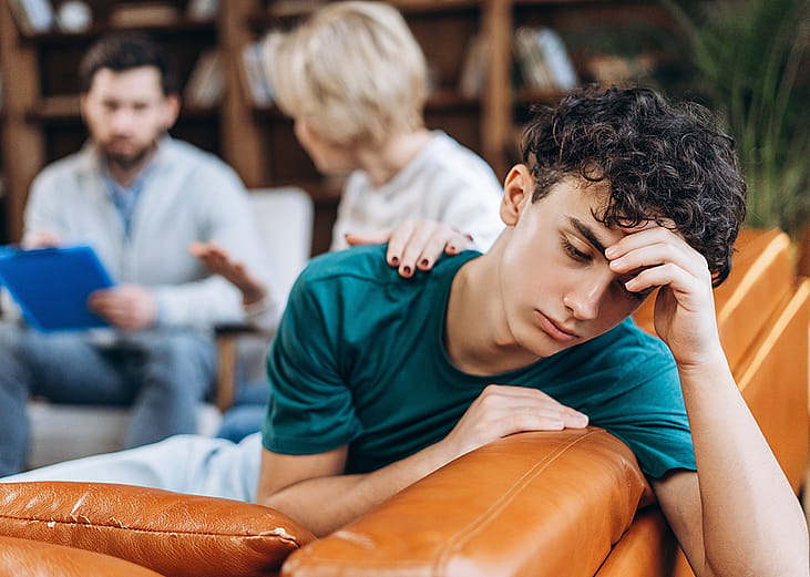 An upset stressed teenage boy waiting for his mother who is talking to a counseling professional.