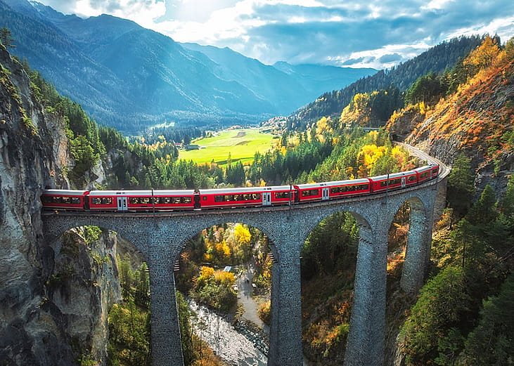 A scenic view of the Bernina Express train on the Landwasser Viaduct with the Alpine mountains and forest during sunset in Switzerland.