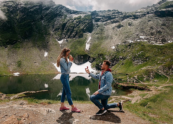 A couple during a marriage proposal with a scenic mountain lake view in the background.