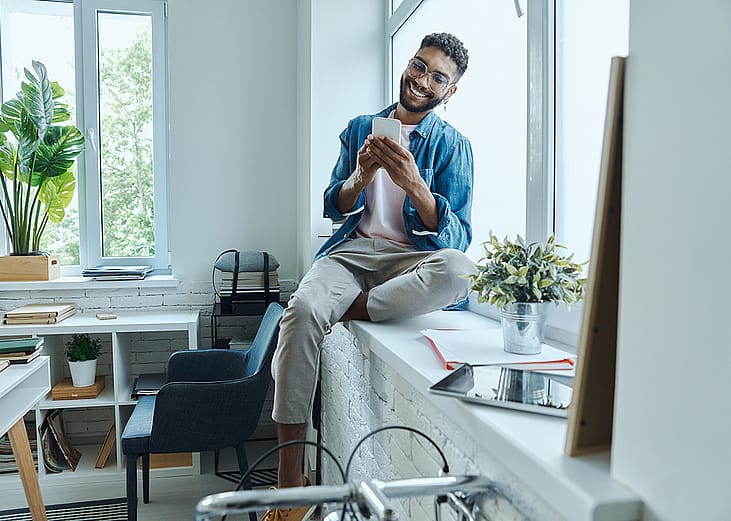 A cheerful man at home using his phone while sitting on the window sill.