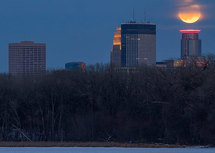 A telephoto shot of the Minneapolis skyscrapers with the full moon disappearing into the twilight sky.