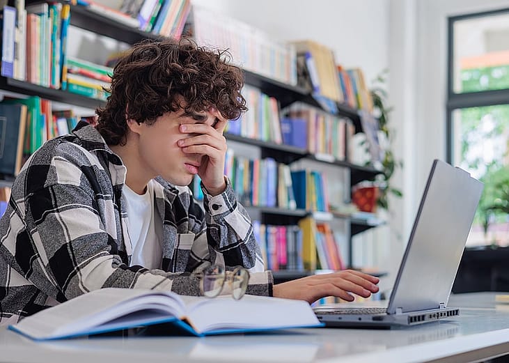 A tired student rubbing his eyes while studying on his laptop in the library.