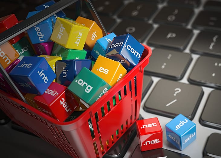A miniature of a shopping basket with colorful blocks of different internet domain extensions placed on top of a laptop's keyboard.