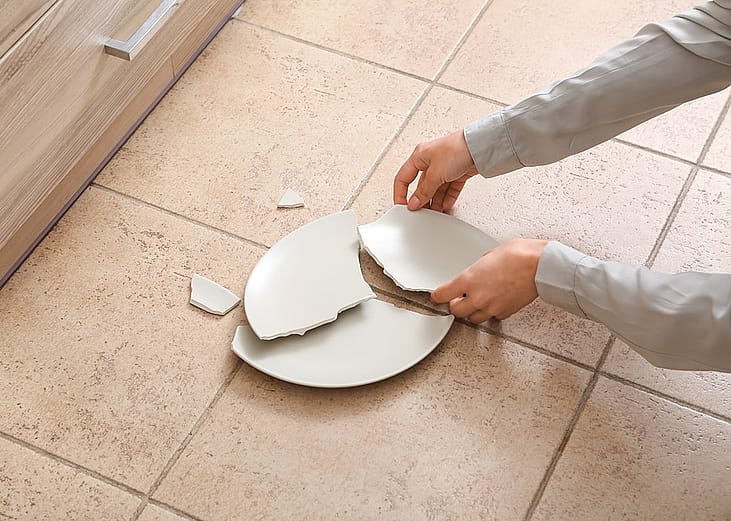 A person picking up a broken ceramic plate on the kitchen floor.