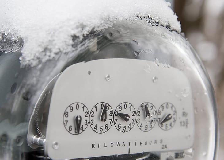 An electric utility meter with snow ice on the top of it.
