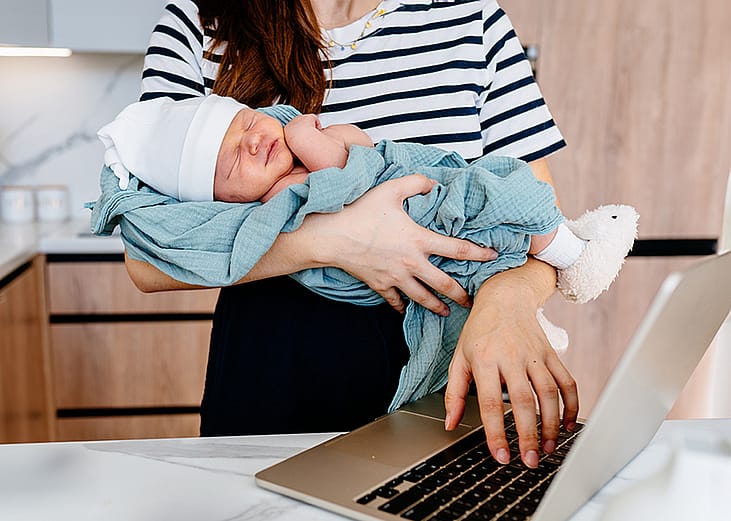 A parent cradling a newborn baby while working remotely from their home kitchen.