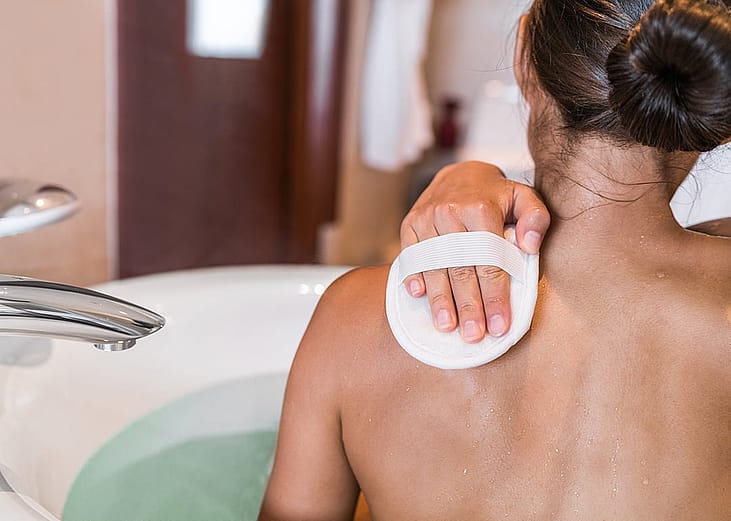 A person exfoliating her back with a scrub sponge in a bathtub.
