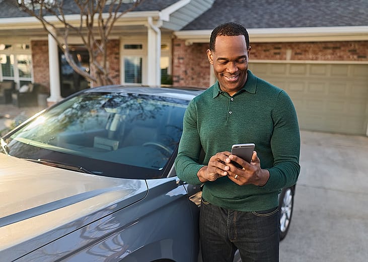 A car owner standing next to his gray car while checking car loans online on a smartphone.