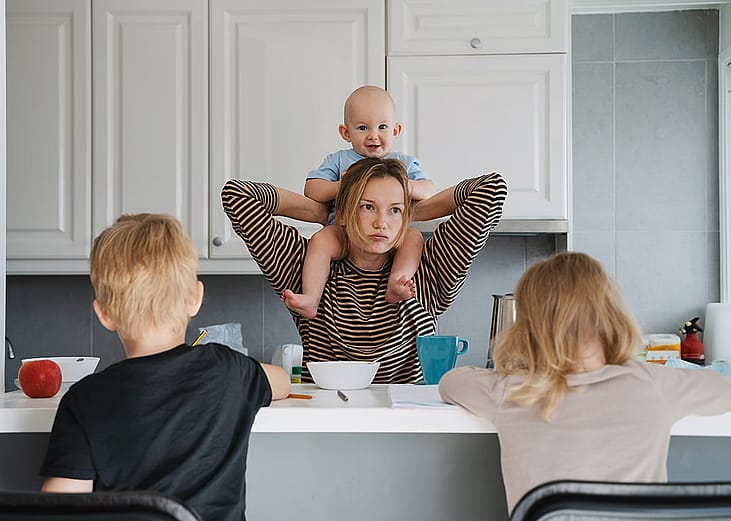 A parent carrying her baby on her shoulders with her other children doing school homeworks in the kitchen countertop.