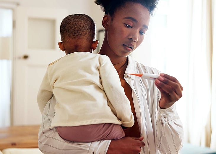 A mother holding her baby and checking temperature with a thermometer.