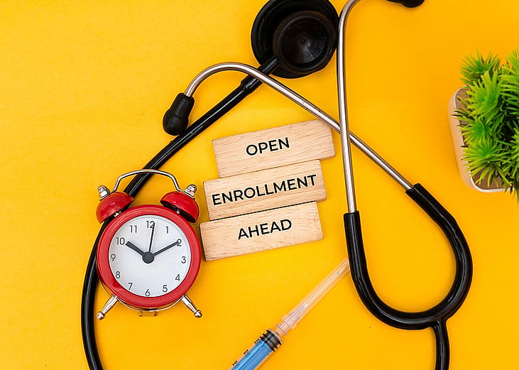 A medical-themed flat lay featuring a stethoscope, a red alarm clock, a syringe, and wooden blocks saying 'Open Enrollment Ahead'.