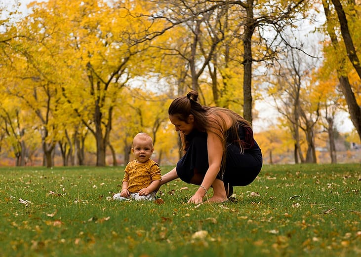 A parent helping her baby pose for a photo in the golden leaves at a park during autumn.