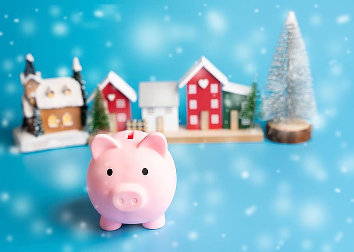 A figure of a piggy bank with Christmas houses and trees in the background as a concept of saving for the holiday season.