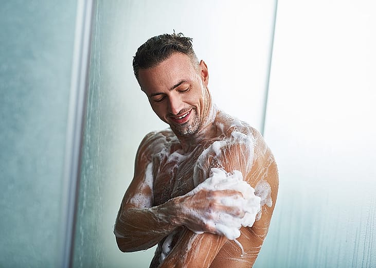 Waist up portrait of a man with foam on his body taking shower at home.