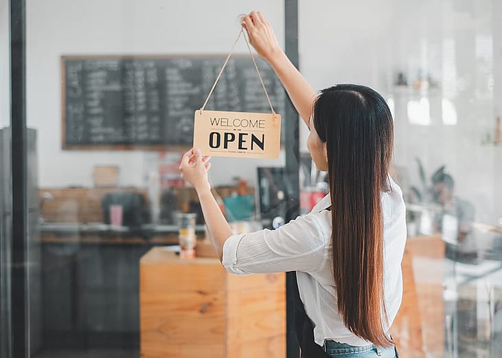 A cafe owner happily puts up a 'Welcome, we are open' sign by the entrance of her shop.