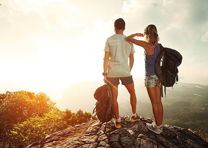 A couple hiking together with backpacks standing on top of a mountain and enjoying valley view.