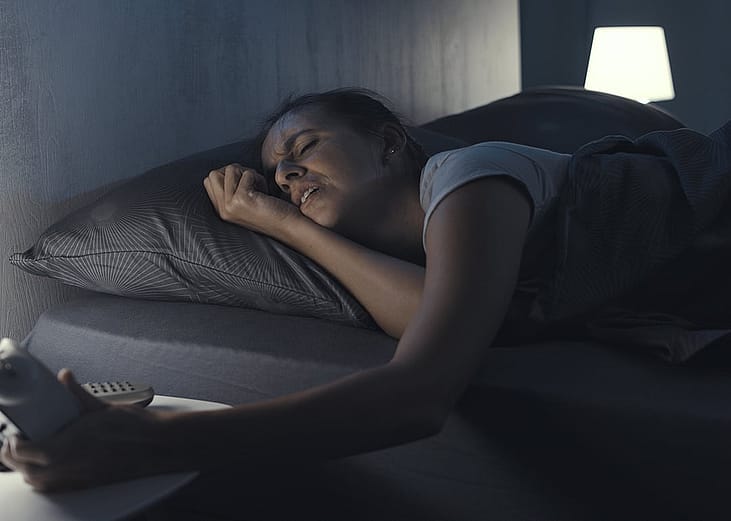A tired woman lying in bed and checking the time on her alarm clock.