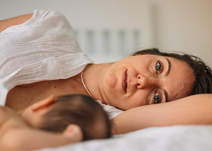 A woman lying on a bed resting next to a newborn baby, looking toward the camera with a tired expression and dark circles under her eyes.