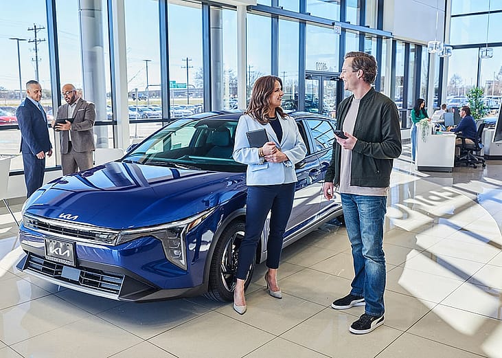 People standing next to a blue Kia sedan inside of a car dealership.