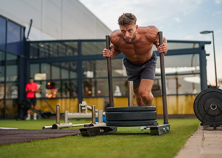 Muscular sportsman performing intense physical exercise with weighted sled on a green turf field in an outdoor gym setting.