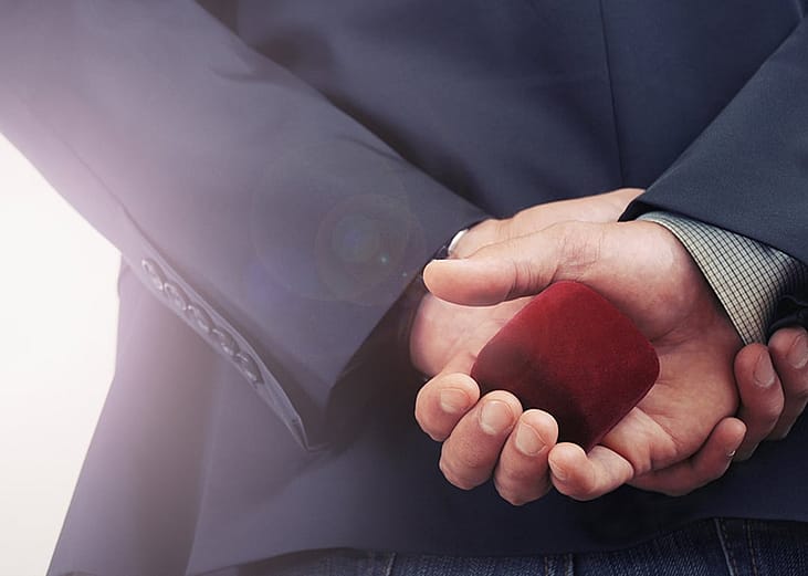 A person holding an engagement ring and its box at his back before a proposal.