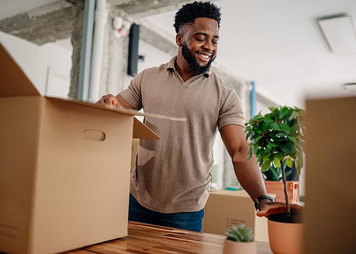 A happy male entrepreneur unpacking boxes and arranging plants while moving into a new office space.