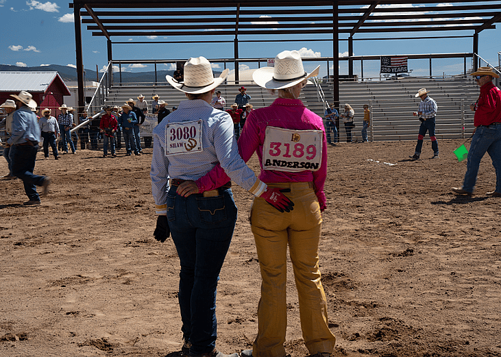 Two rodeo participants stand with their arms around each other's backs looking at the rodeo grounds.
