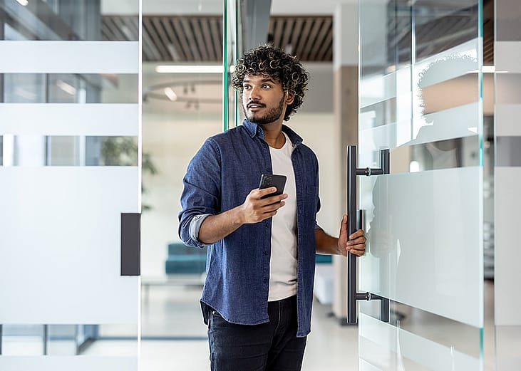 A male employee holding a phone while opening the glass door to an office.