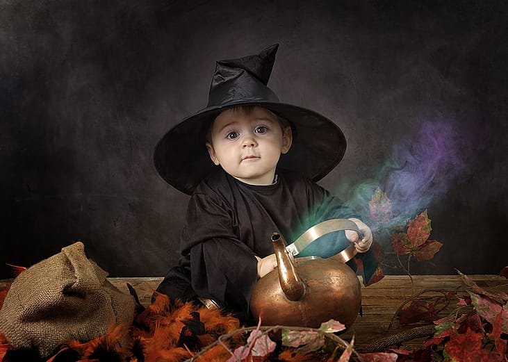 A young baby dressed in a classic black witch costume with a pointed witch hat, sitting beside a copper pot or kettle with colorful smoke or steam rising from it.