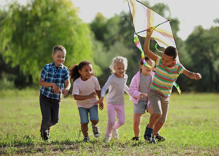 A group of happy kids running in a park. One boy is holding a kite. 