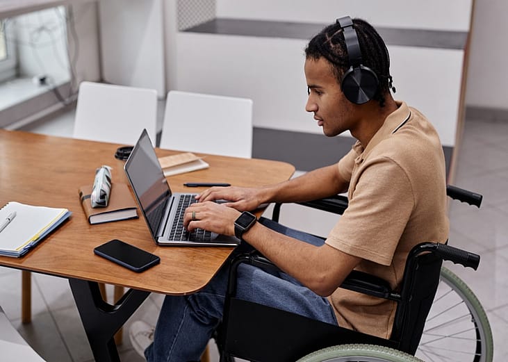 A man in a wheelchair using headphones at a laptop.