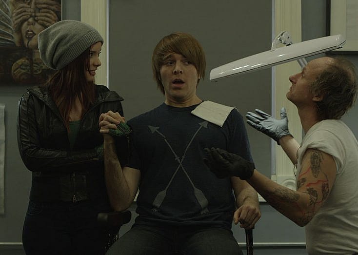 A boy and girl holding hands while a tatoo artist throws his hands in the air.