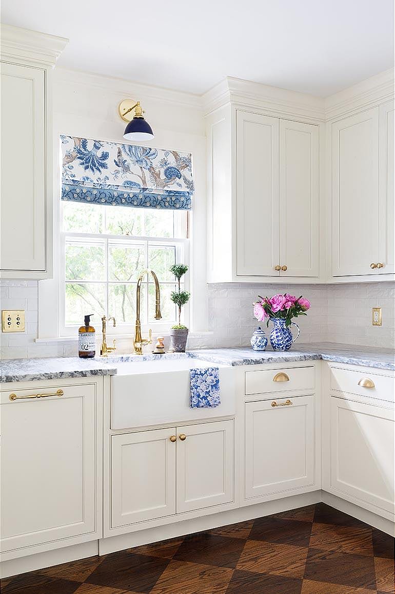 White cabinetry and a golden faucet with a window curtain that is blue and white like the wallpaper.