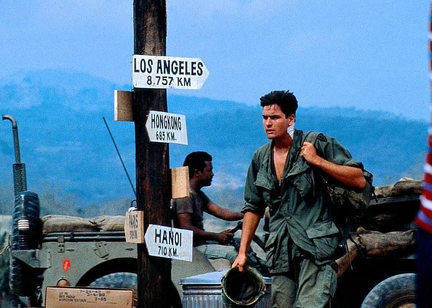Charlie Sheen in military gear walking past a sign pointing to various cities with a military jeep in the background. 
