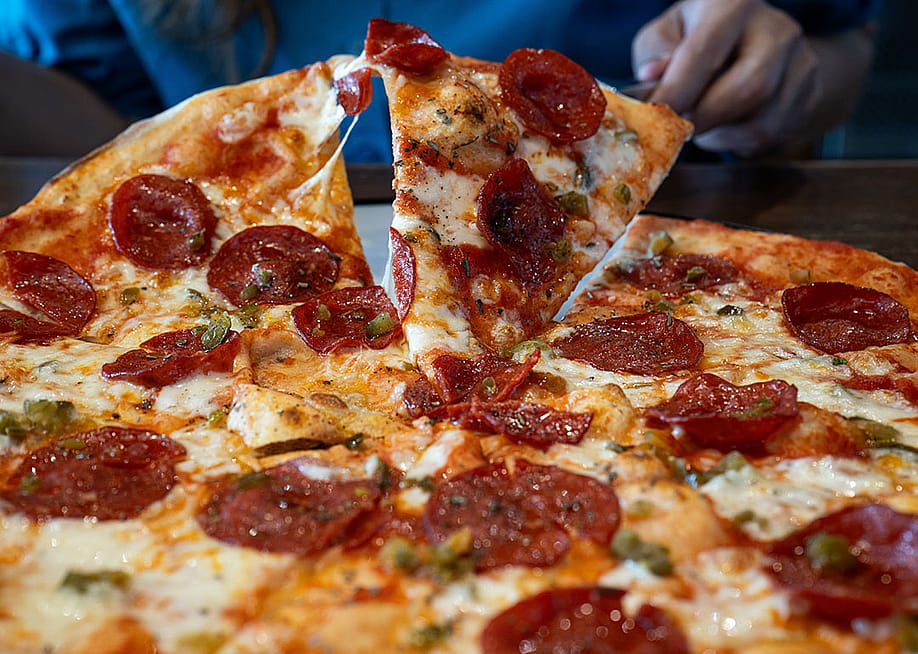 Slice of appetizing pepperoni pizza with melting cheese is being up and picked up from whole pizza by a person off screen. — joebarthez // Shutterstock.