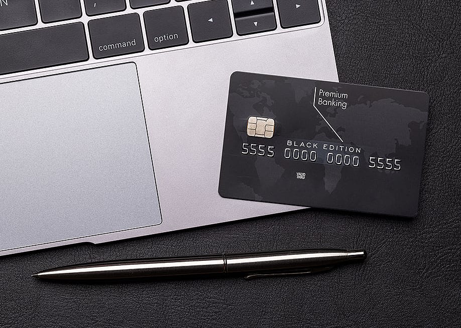 Black premium bank card lays on a silver macbook laptop computer next to the trackpad, with a shiny fancy pen on the table. — evgeeenius // Shutterstock