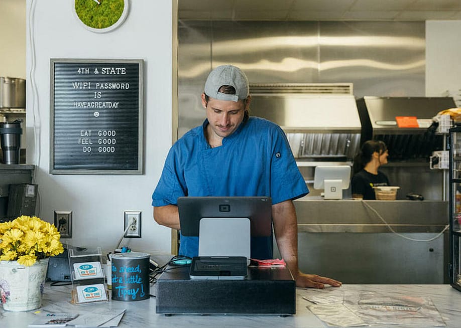 Person wearing a blue shirt entering items into a tablet cash register behind the counter with a kitchen and sign int he background. — Square