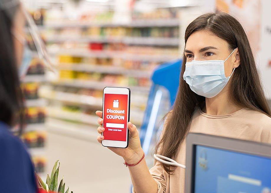 Woman holding up a smart phone with red discount coupon displayed with and drugstore shelves in the background. — Pressmaster // Shutterstock