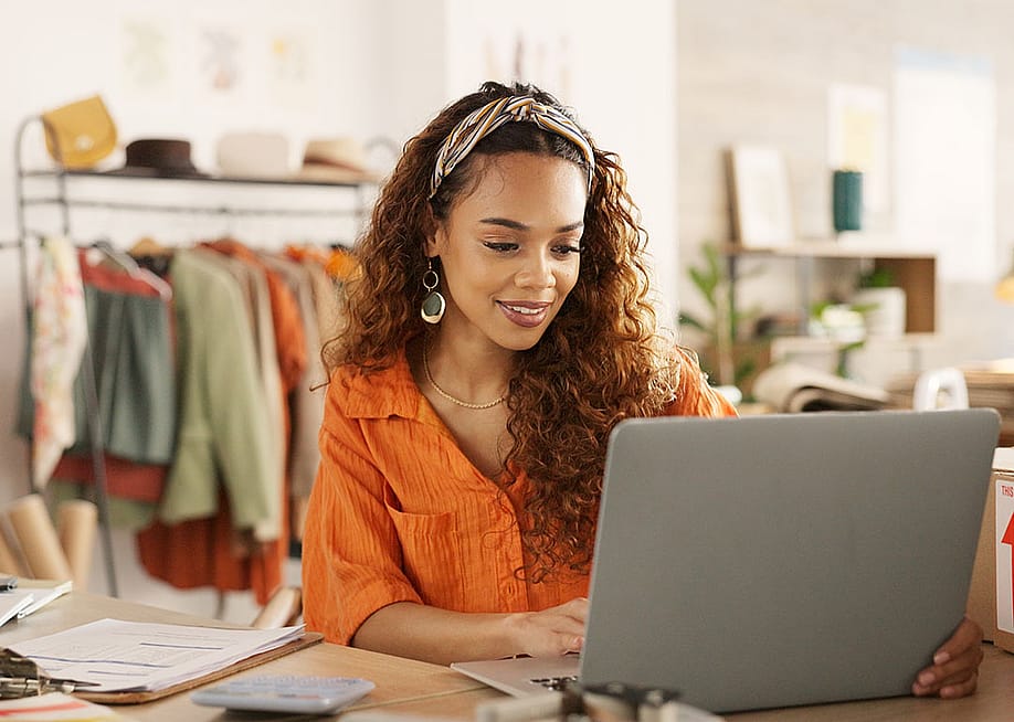 Black woman using laptop in clothing store working on business with rolling rack of clothes behind her. — Yuri A. // Shutterstock