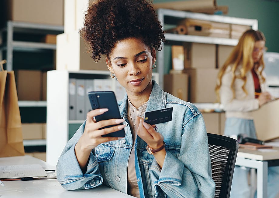Black woman entrepreneur looking at a business credit card and holding smartphone inside an office with boxes and shelves and a blurred blond person in the background. — Yuri A. // Shutterstock