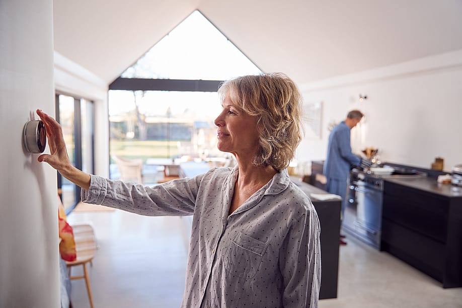 Middle-aged woman wearing pajamas adjusts modern thermostat in a bright house with a vaulted ceiling while a person wearing blue pajamas is seen in the kitchen counter in the background. — Monkey Business Images // Shutterstock