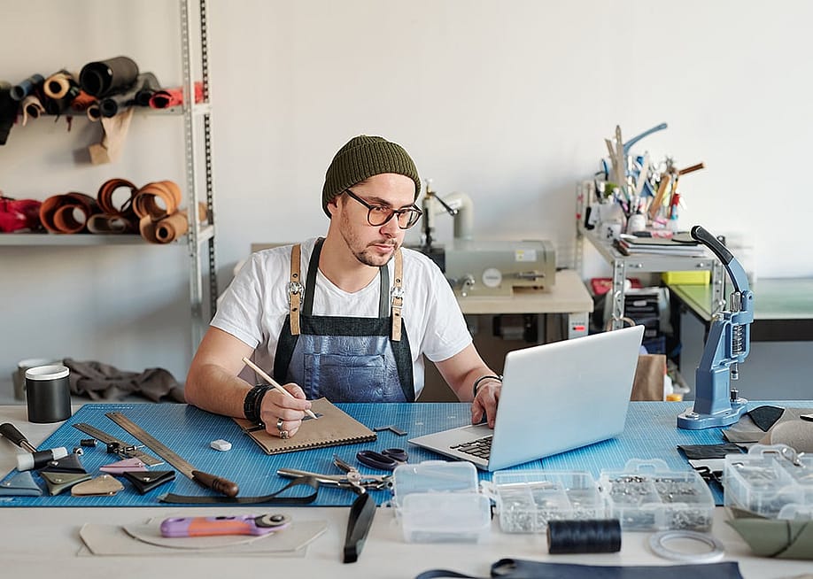 A leather goods craftsman using a laptop and notebook to start his production process. — Pressmaster // Shutterstock