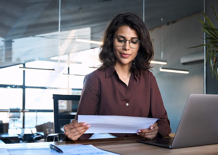 Focused young businesswoman reading financial report document and using a laptop in the workplace. — Stock 4you // Shutterstock