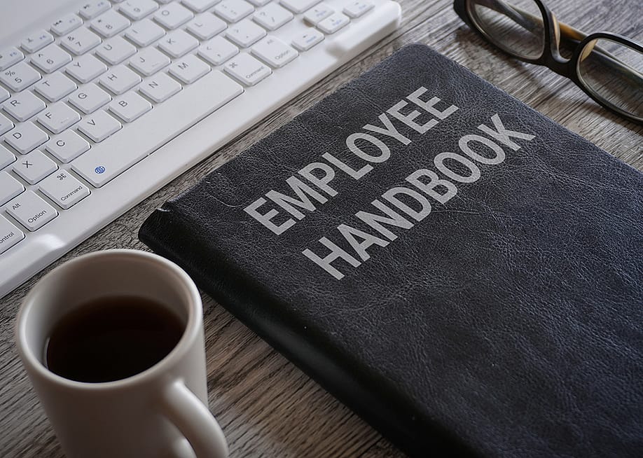 An employee handbook and a cup of coffee in front a work computer keyboard. — izzuanroslan // Shutterstock